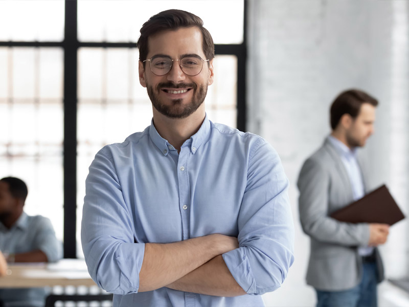 Smiling man with glasses in a blue shirt stands confidently in a modern office environment.