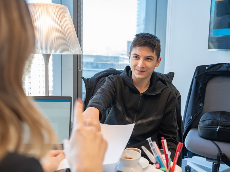 A young person shaking hands in an office setting near a window.