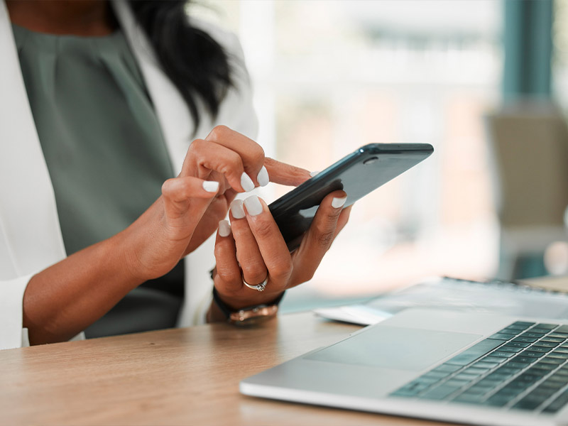 A woman uses a smartphone at a desk with a laptop, focusing on technology and communication.