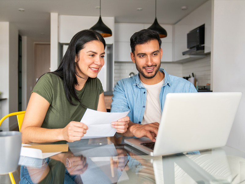 A couple smiling and using a laptop at home while checking documents at a table.