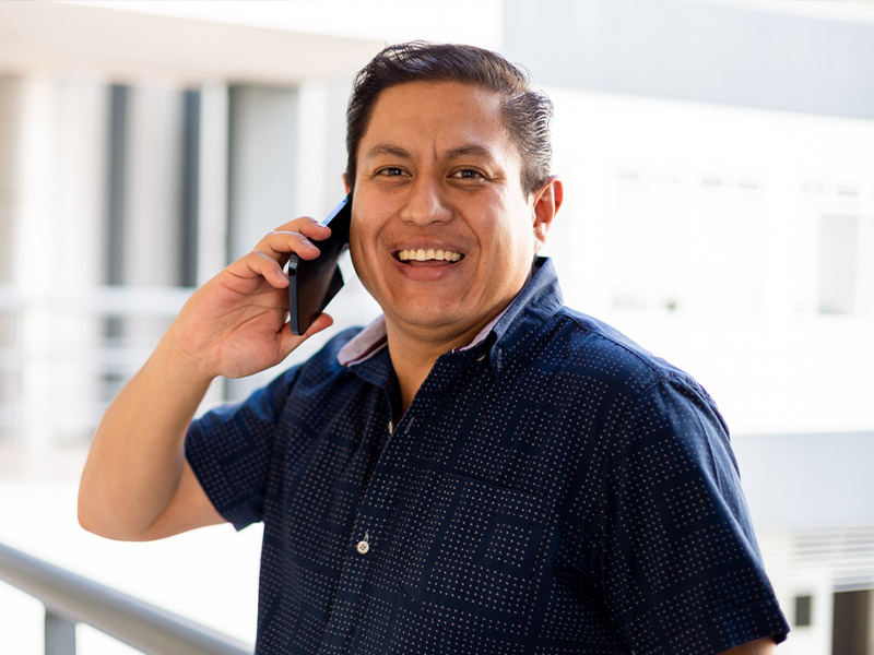 A man smiling while talking on a smartphone, standing in a bright indoor setting.