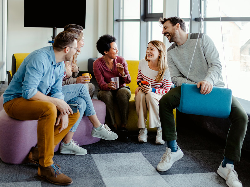 Group of people sitting on colorful seats, laughing and talking in a modern office setting.