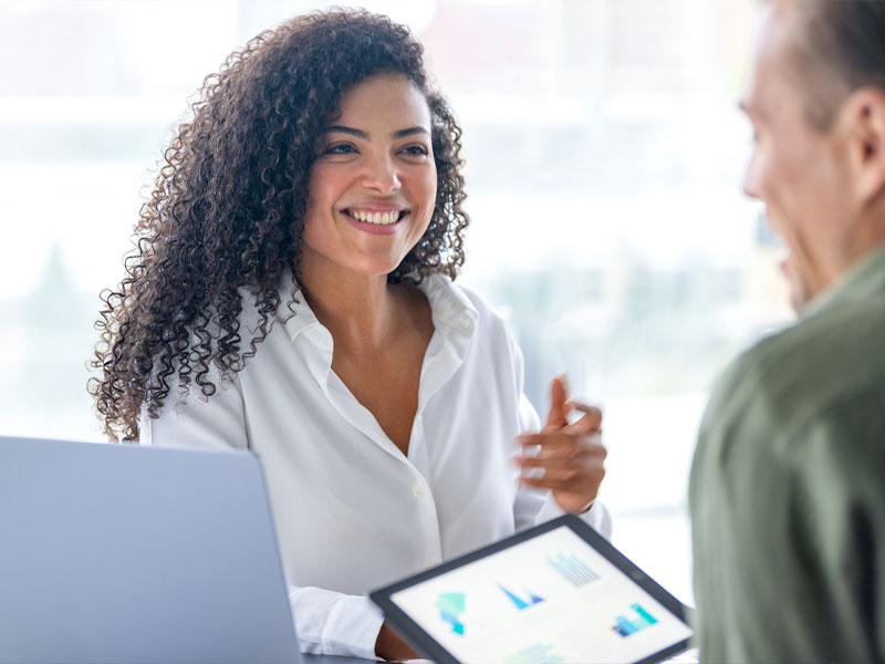 A woman smiling in a business meeting with a colleague, holding a tablet displaying charts.