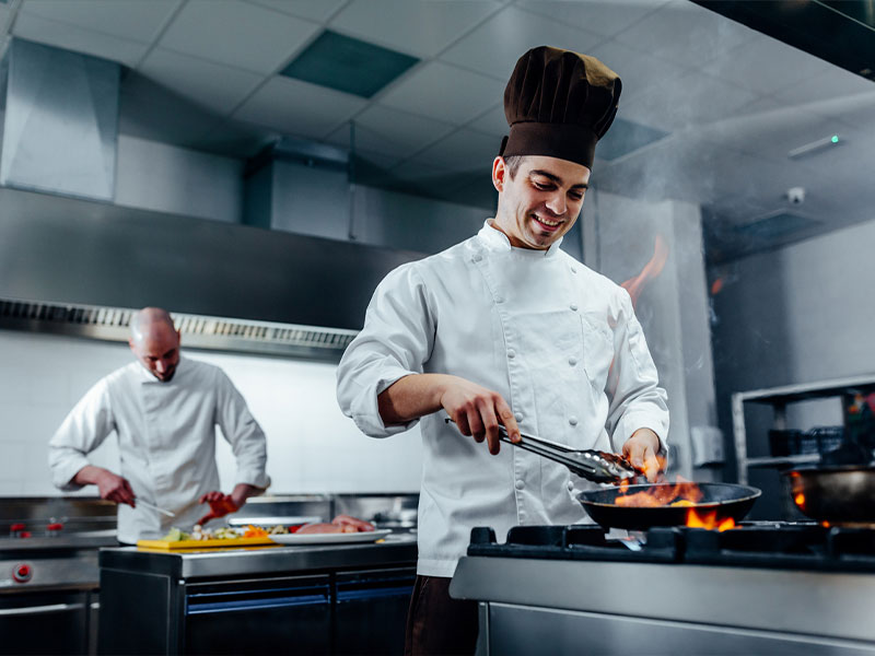 Chef smiling while cooking with flames in a professional kitchen.