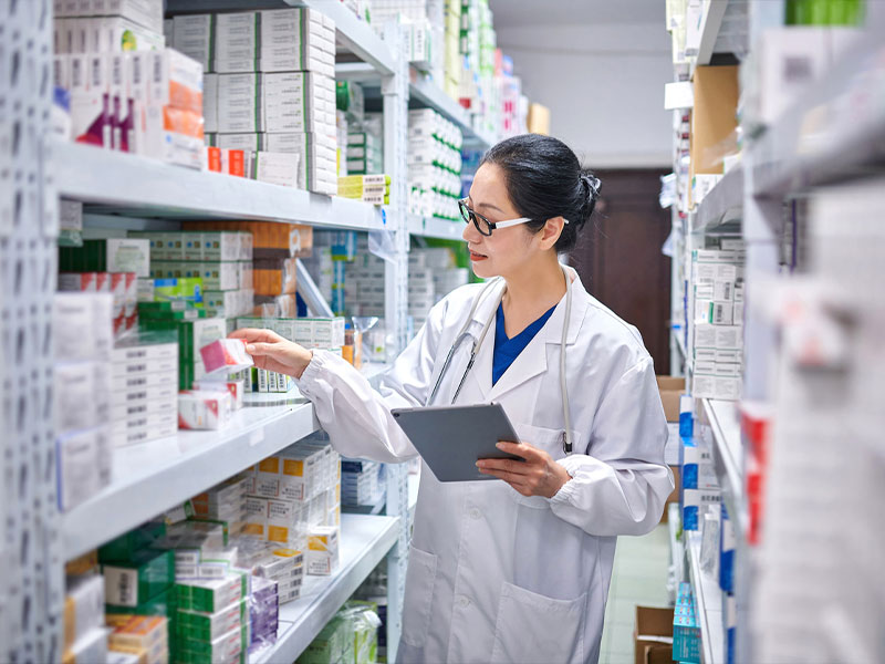 Pharmacist checking medication stock on shelves, holding a tablet in a pharmacy.