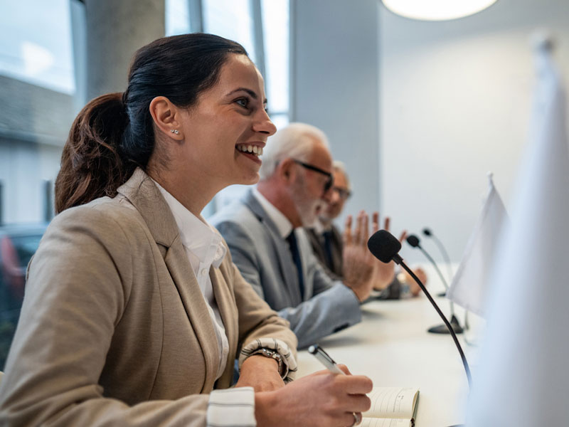 Smiling woman at conference table with colleagues and microphones, taking notes.