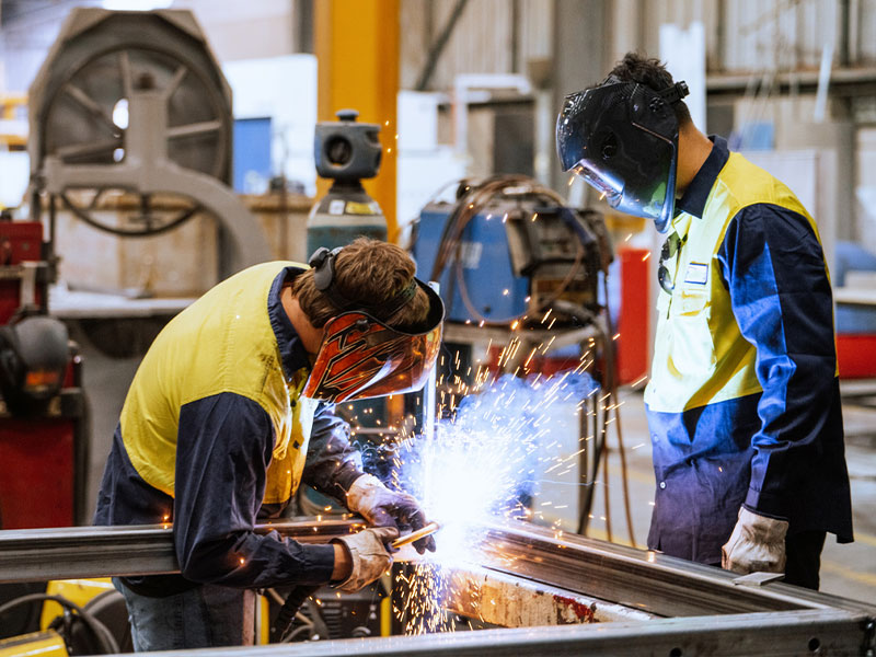 Two workers in a factory welding metal, wearing protective helmets and yellow safety vests.