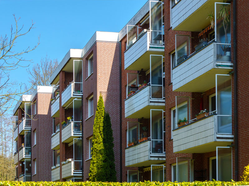 Red-brick apartment building with balconies, surrounded by trees on a sunny day.