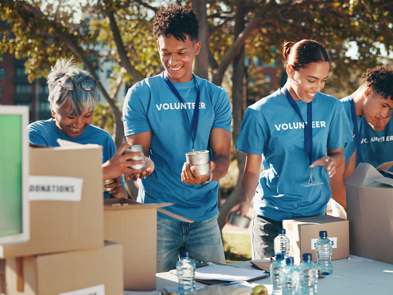 Volunteers in blue shirts packing donation boxes outdoors with canned goods and bottles.