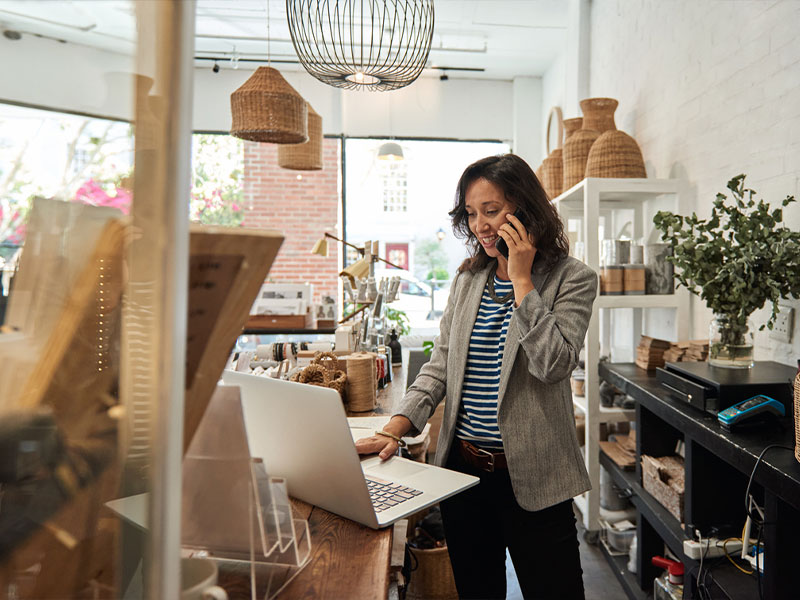 Woman in a shop talking on phone and using a laptop.