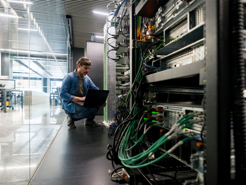 Person working on a laptop in a server room with network cables and servers.