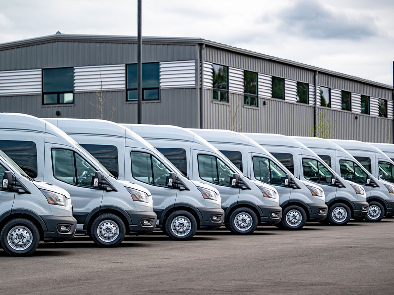 Row of silver vans parked outside a large industrial building on a clear day.