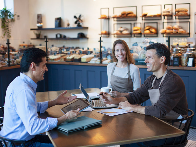 Three people meeting in a bakery, discussing business plans over coffee and laptops.