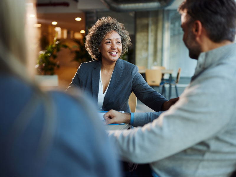 A woman in a gray blazer smiling while engaged in conversation with two people in an office setting.