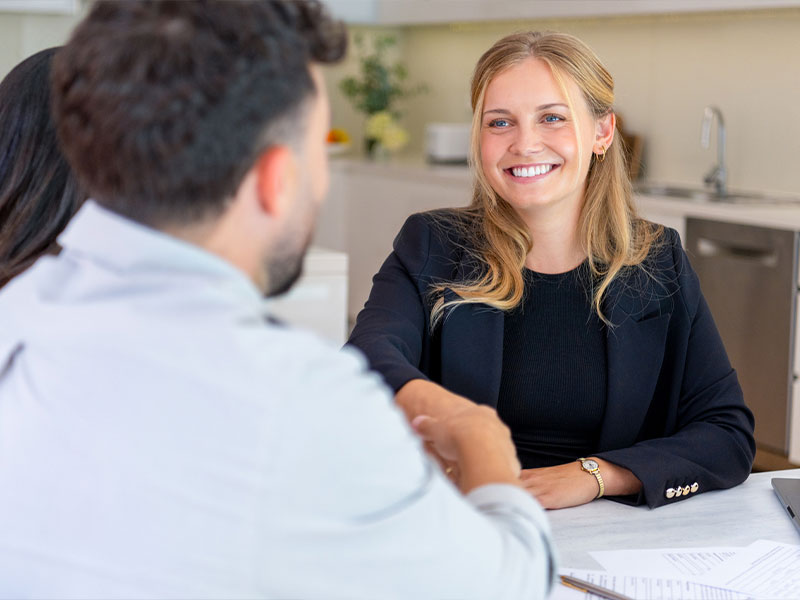 Woman in a blazer smiling and shaking hands with a man in a business setting.