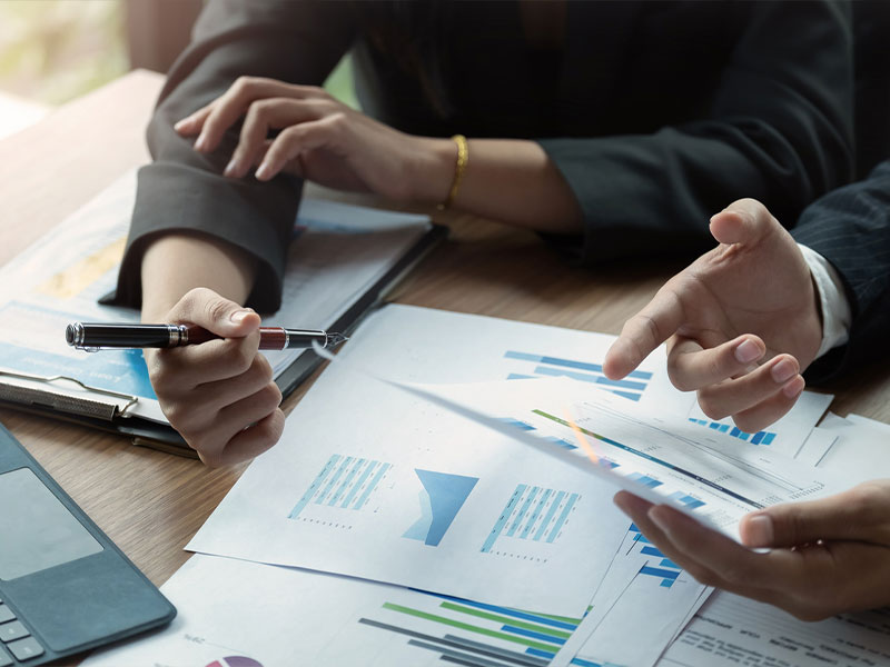 Two people discussing documents at a meeting, charts and graphs visible on the table.