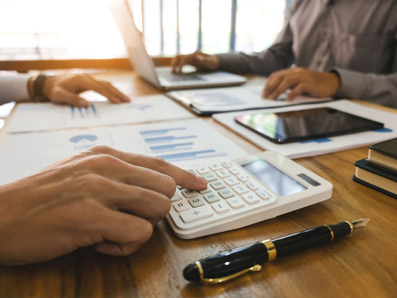 Hands using a calculator on a desk with charts, laptop, and tablet.