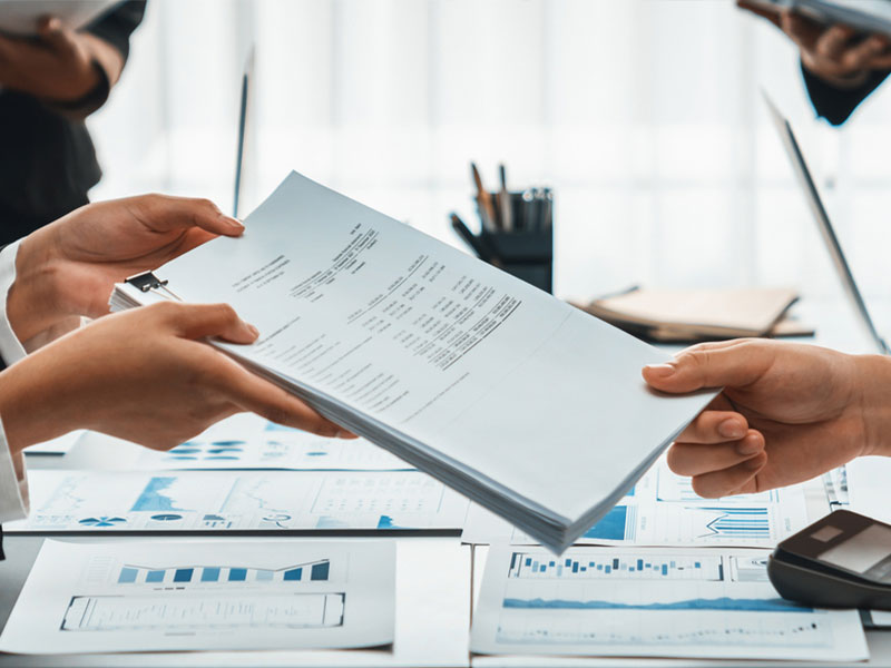 Two people exchanging business documents over a desk with charts.