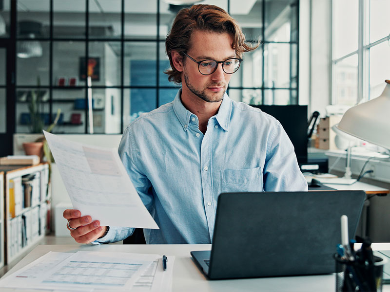 A man in a blue shirt reviews documents while working on a laptop in a modern office.