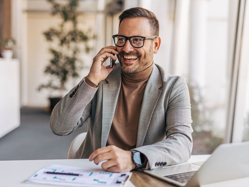 A man in a blazer smiling while talking on a phone in an office setting.
