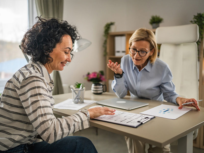 Two women discussing paperwork at a desk in a bright office environment.