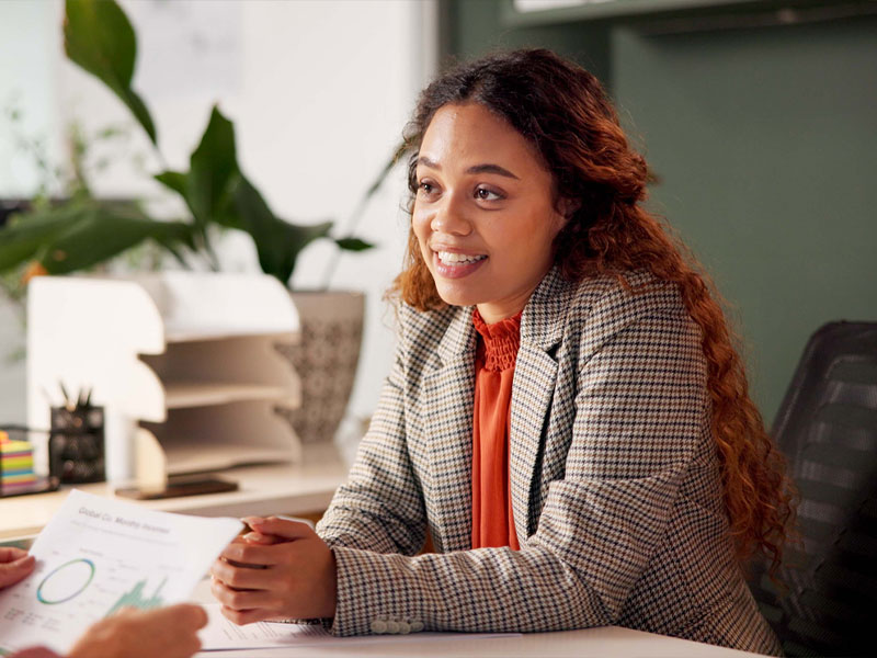 A woman in an office setting discussing a document, surrounded by plants and office supplies.
