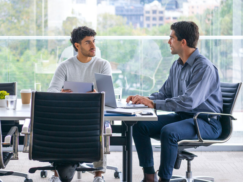 Two men having a discussion at a table with laptops in a bright office setting.