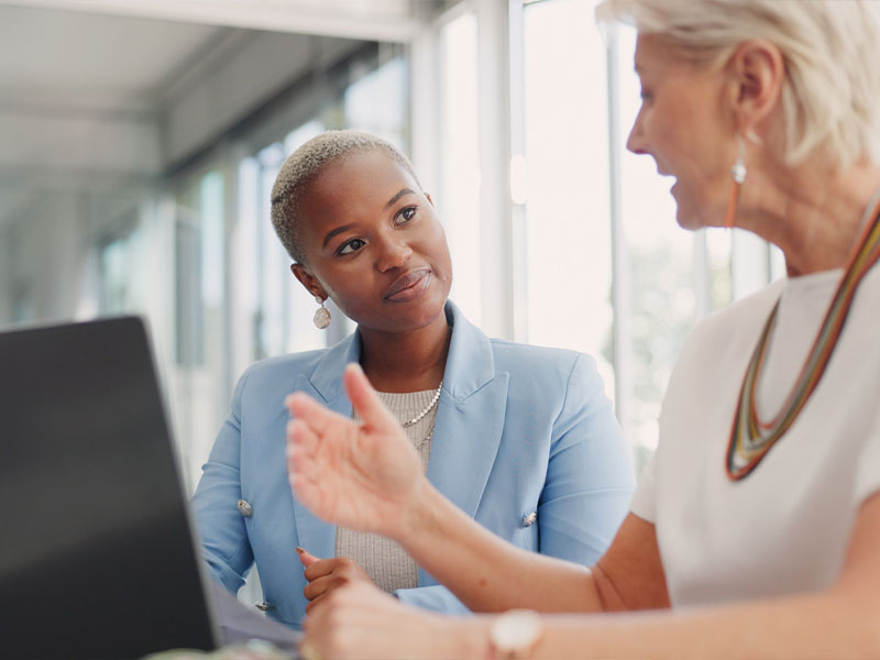 Two women in a business meeting, one attentively listening while the other speaks, next to a laptop in an office setting.
