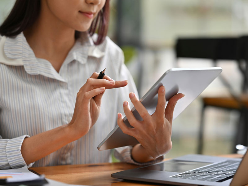 A woman using a tablet and holding a stylus at a desk with a laptop.