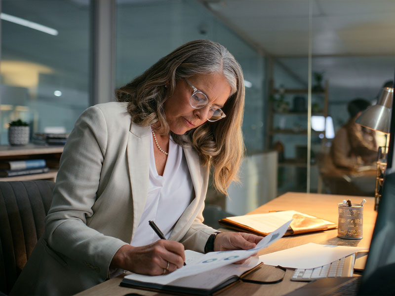 A woman in a blazer working at a desk, writing notes in a notebook, in a modern office setting.