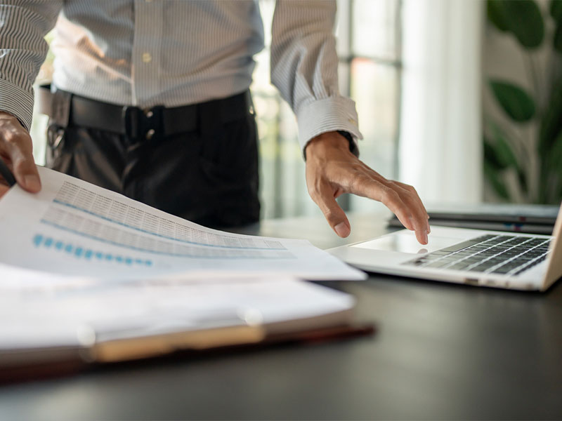 A person working at a desk, reviewing charts on paper and using a laptop in a bright office.