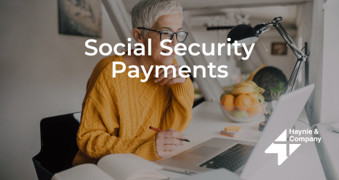 Senior person in sweater working on laptop at a desk labeled Social Security Payments with Haynie logo.