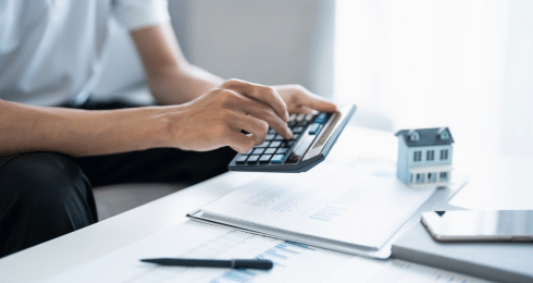 Person using calculator with small house model on desk, managing finances.