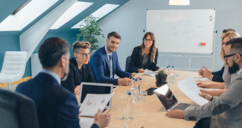 A group of business professionals engaged in a meeting around a conference table with laptops and documents.