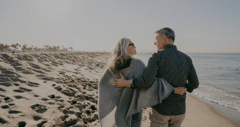 Elderly couple walking arm in arm on a sunny beach.