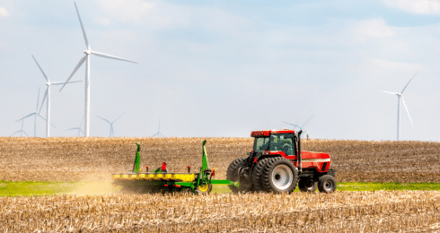 Red tractor plowing a field with wind turbines in the background under a blue sky.