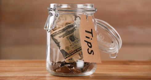A glass jar labeled Tips filled with coins and a dollar bill on a wooden table.