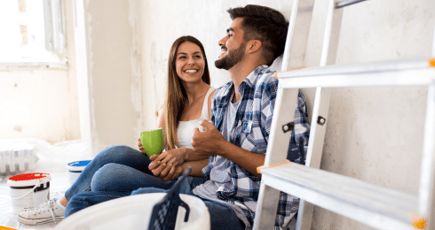 Couple smiling and relaxing during a home renovation, sitting on the floor with paint supplies nearby.