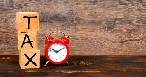 Wooden blocks spelling TAX next to a red alarm clock on a wooden surface.