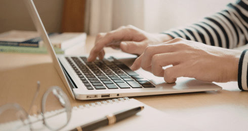 Person typing on a laptop at a desk, with glasses and a notepad nearby.