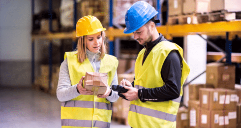 Two warehouse workers in safety vests and helmets checking inventory.