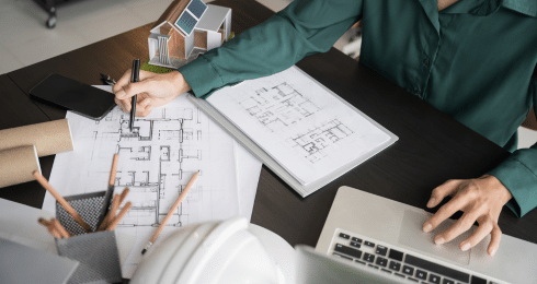 Architect working on blueprints with a laptop and model house on the desk.