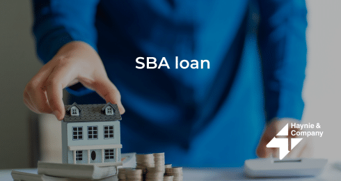 A person in blue placing miniature house by coin stacks, text SBA loan, Haynie logo.