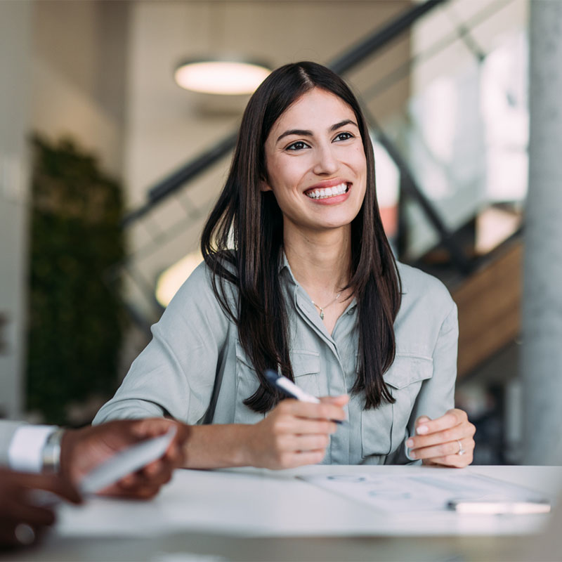 A woman smiling while writing at a desk in an office setting.