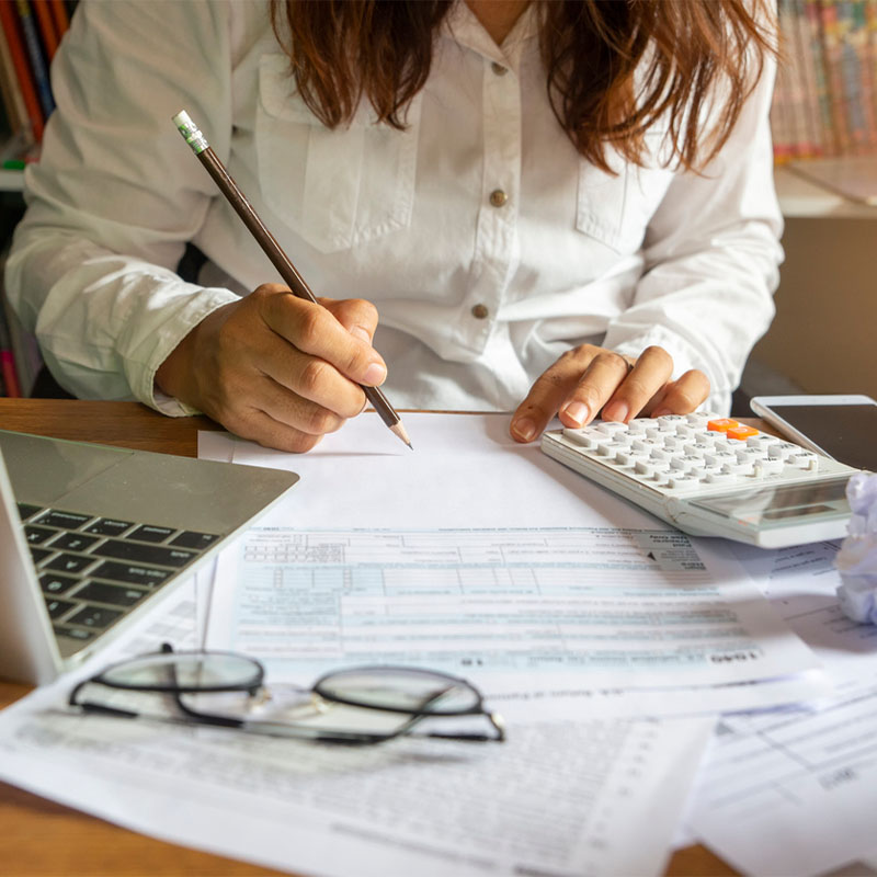 Woman in white shirt using calculator and pencil for financial paperwork by laptop and glasses.