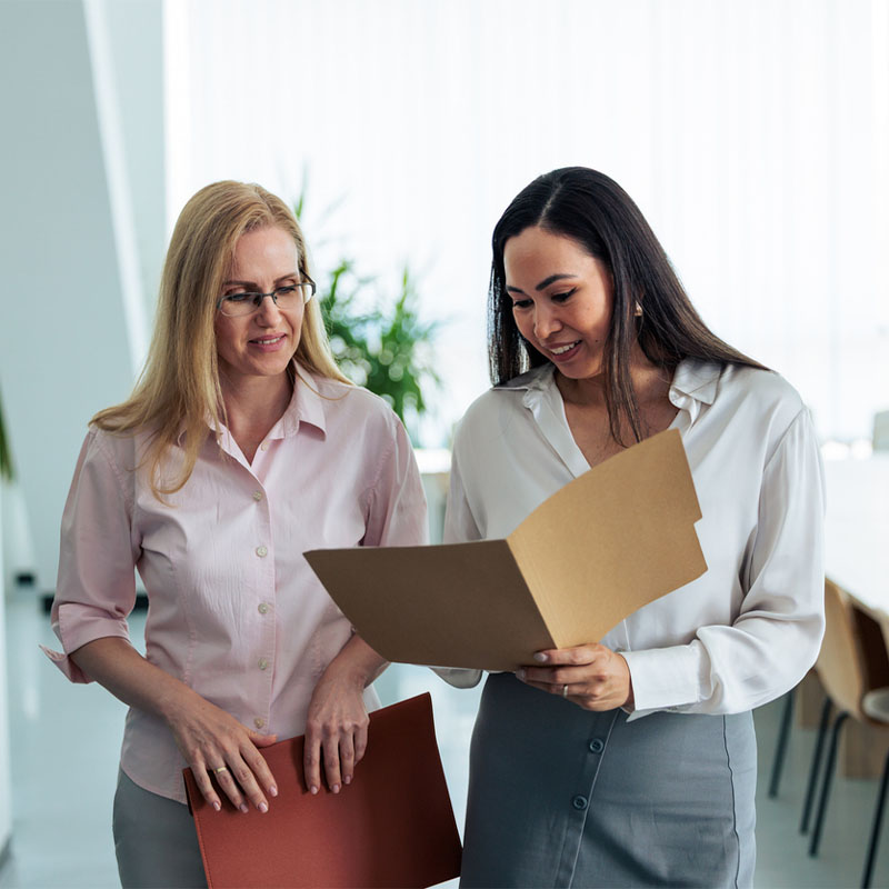 Two women in an office reviewing documents together, one holding a folder.