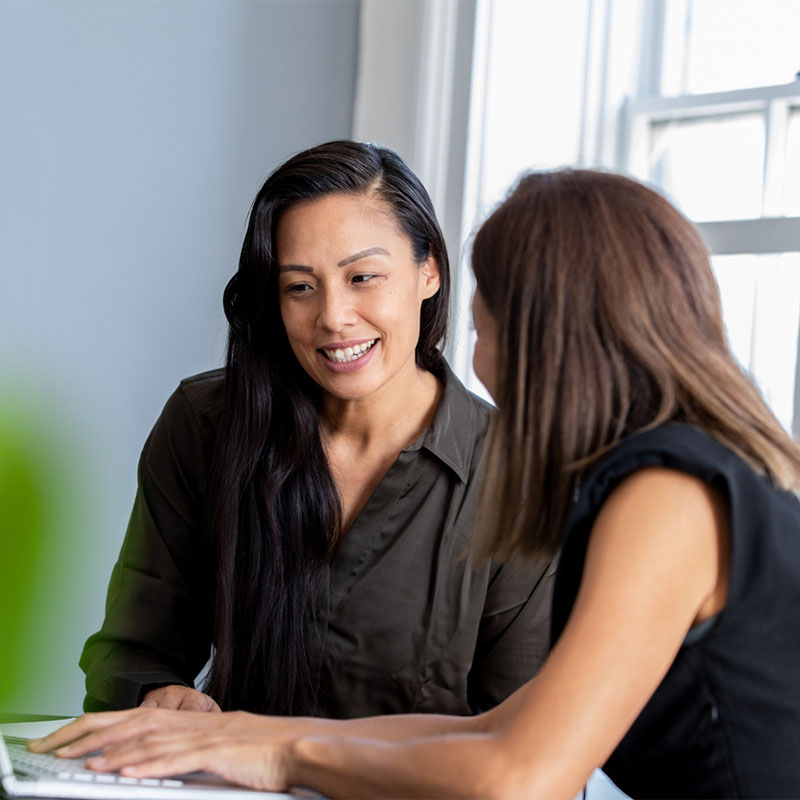 Two women discussing work at a desk with a laptop in a bright, windowed room.
