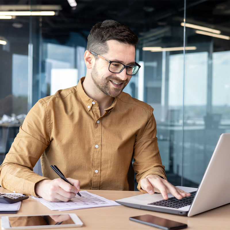 Man in brown shirt working on laptop, smiling in modern office setting.