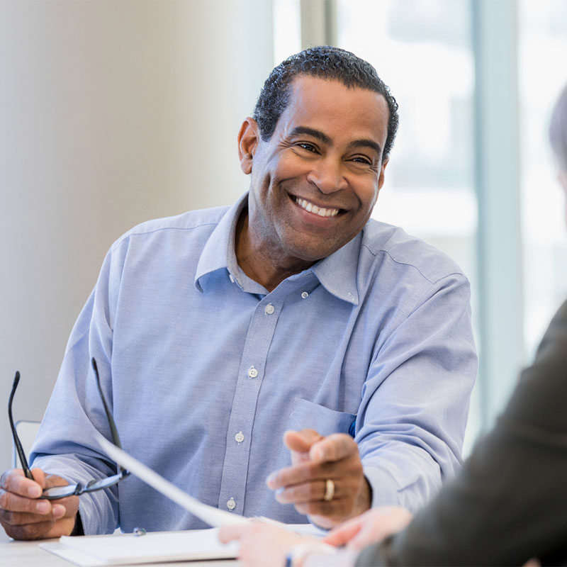 Smiling man in a blue shirt engaging in a meeting, holding glasses and a pen.