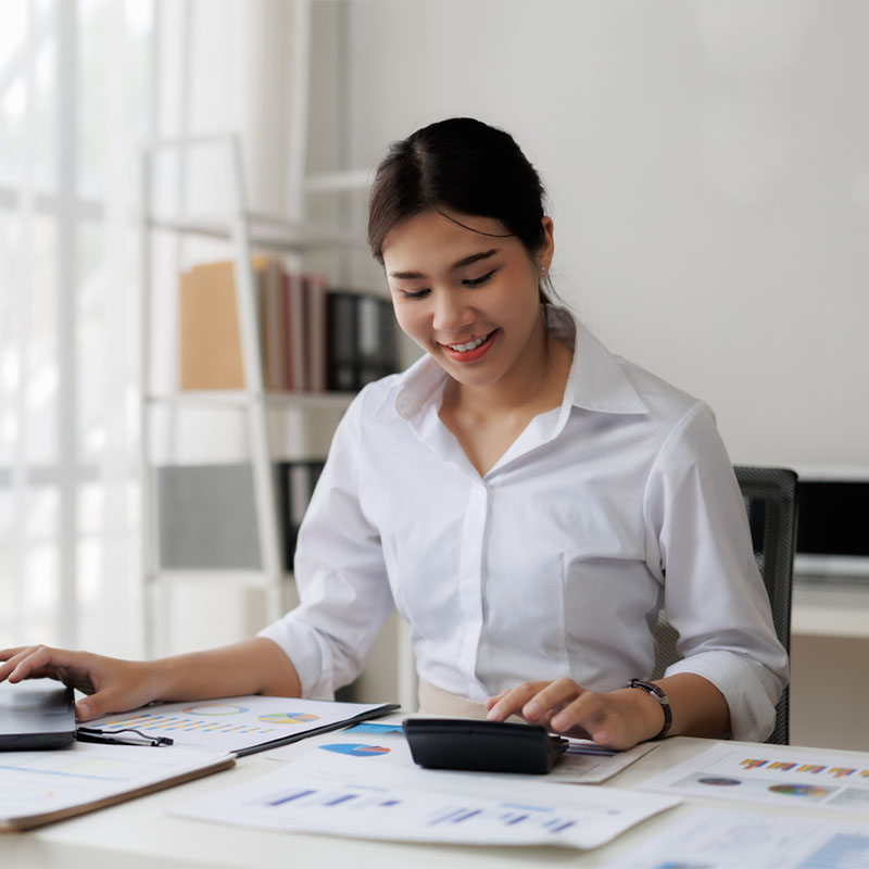 A woman in a white shirt using a calculator at a desk with documents in an office.